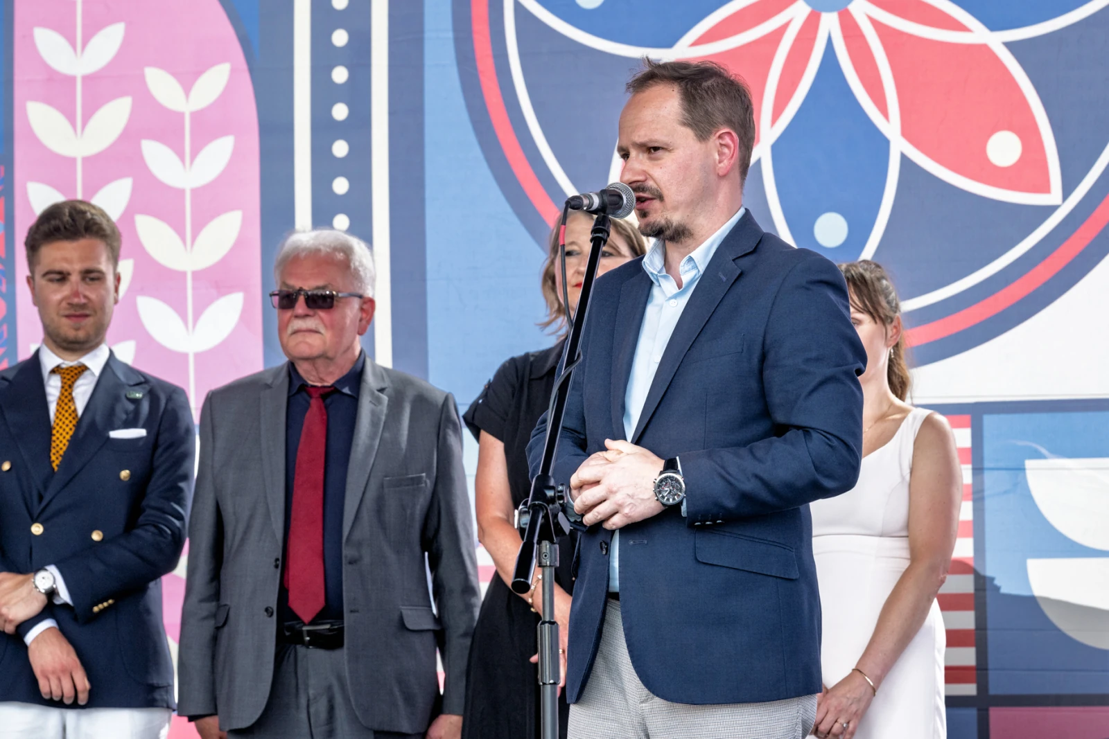 Mężczyzna w garniturze przemawia do mikrofonu, obok stoją inni uczestnicy wydarzenia.  Autor: Piotr Wojnarowski