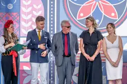 Grupa osób w tradycyjnych i eleganckich strojach, stojących obok siebie na tle kolorowych wzorów.
