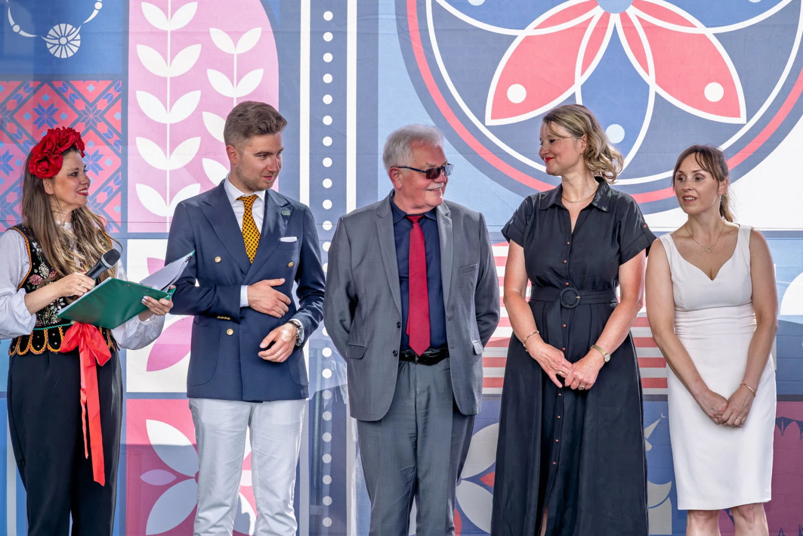 Grupa osób w tradycyjnych i eleganckich strojach, stojących obok siebie na tle kolorowych wzorów.  Autor: Piotr Wojnarowski