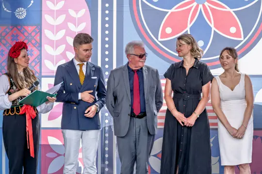 Grupa osób w tradycyjnych i eleganckich strojach, stojących obok siebie na tle kolorowych wzorów.