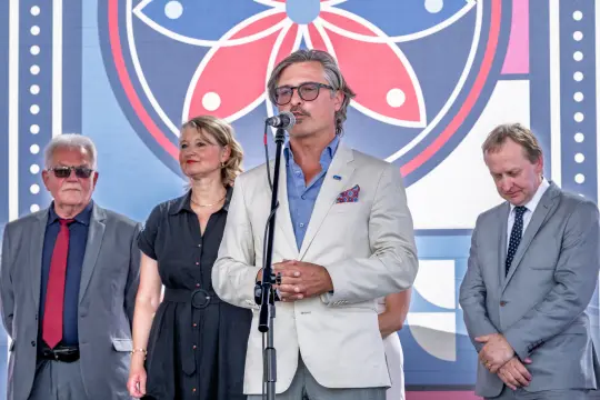 Mężczyzna w jasnym garniturze przemawia do mikrofonu, obok stoją trzy osoby.
