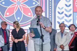 Mężczyzna z mikrofonem przemawia na tle kolorowej dekoracji, obok niego stoi grupa osób.