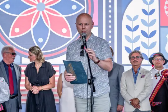 Mężczyzna z mikrofonem przemawia na tle kolorowej dekoracji, obok niego stoi grupa osób.