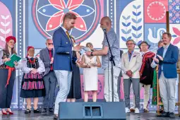 Mężczyzna w garniturze odbiera nagrodę na tle kolorowej dekoracji, wśród innych osób w tradycyjnych strojach.