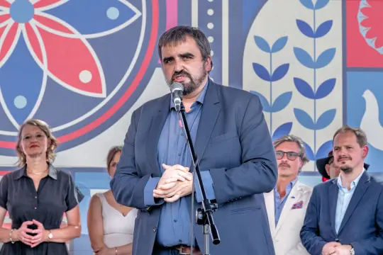 Mężczyzna w garniturze przemawia do mikrofonu, w tle grupa osób.