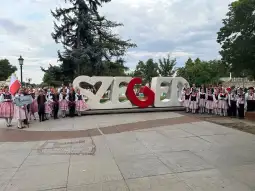 Grupa ludzi w tradycyjnych strojach ludowych stoi przy dużym napisie "Szeged".