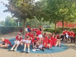 Grupa dzieci w czerwonych koszulkach siedzi na trawie, czytając książki w parku.