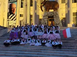 Grupa dzieci w tradycyjnych strojach ludowych z flagą Polski przed budynkiem.