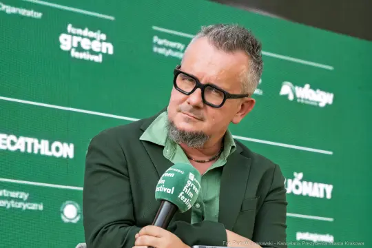 Mężczyzna w okularach, w zielonej koszuli i marynarce, rozmawia podczas festiwalu.