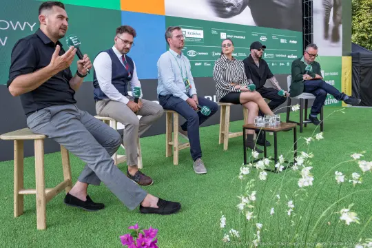 Panel dyskusyjny z sześcioma mężczyznami siedzącymi na stołkach, w tle zielona scena i kwiaty.