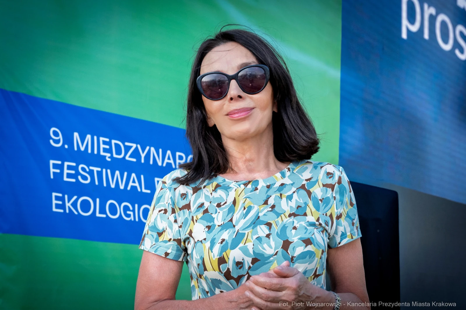 Kobieta w okularach przeciwsłonecznych, ubrana w kolorową sukienkę, stoi przed tłem festiwalu ekologicznego.  Autor: Piotr Wojnarowski