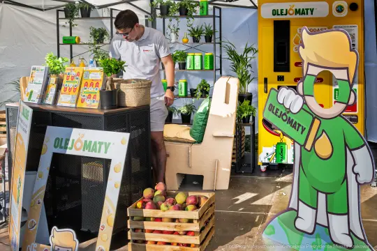 Stoiska promocyjne z roślinami, produktami i postacią maskotki.