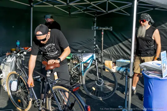 Mężczyzna naprawia rower pod namiotem, obok stoi drugi mężczyzna z brodą.