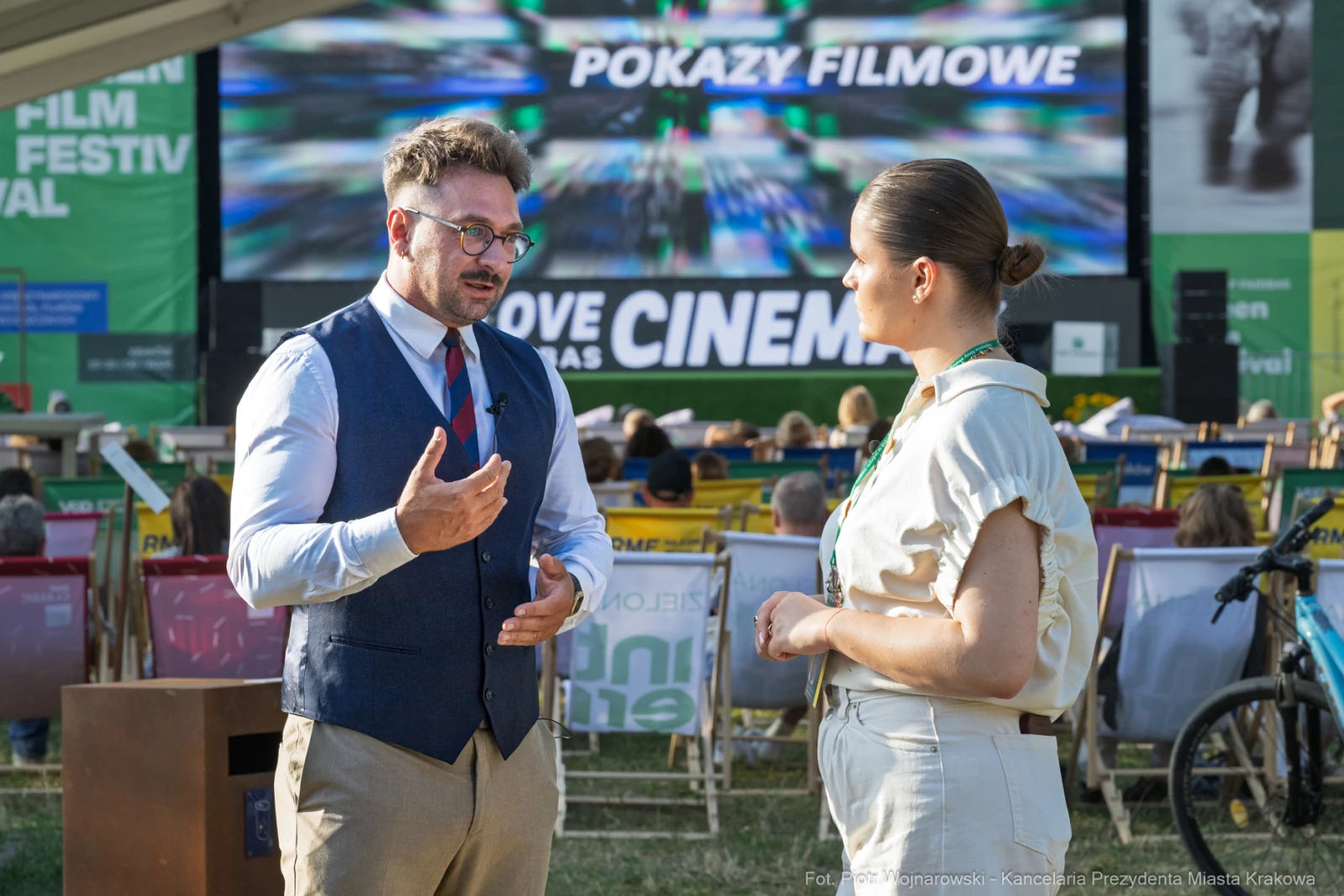 Dwie osoby rozmawiają na tle ekranu z pokazami filmowymi podczas festiwalu.  Autor: Piotr Wojnarowski