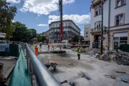 Prace budowlane na ulicy, dźwig podnoszący elementy konstrukcyjne, robotnicy w odzieży ochronnej.