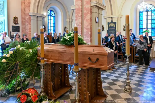 Trumna w kaplicy otoczona przez zgromadzonych ludzi i kwiaty.