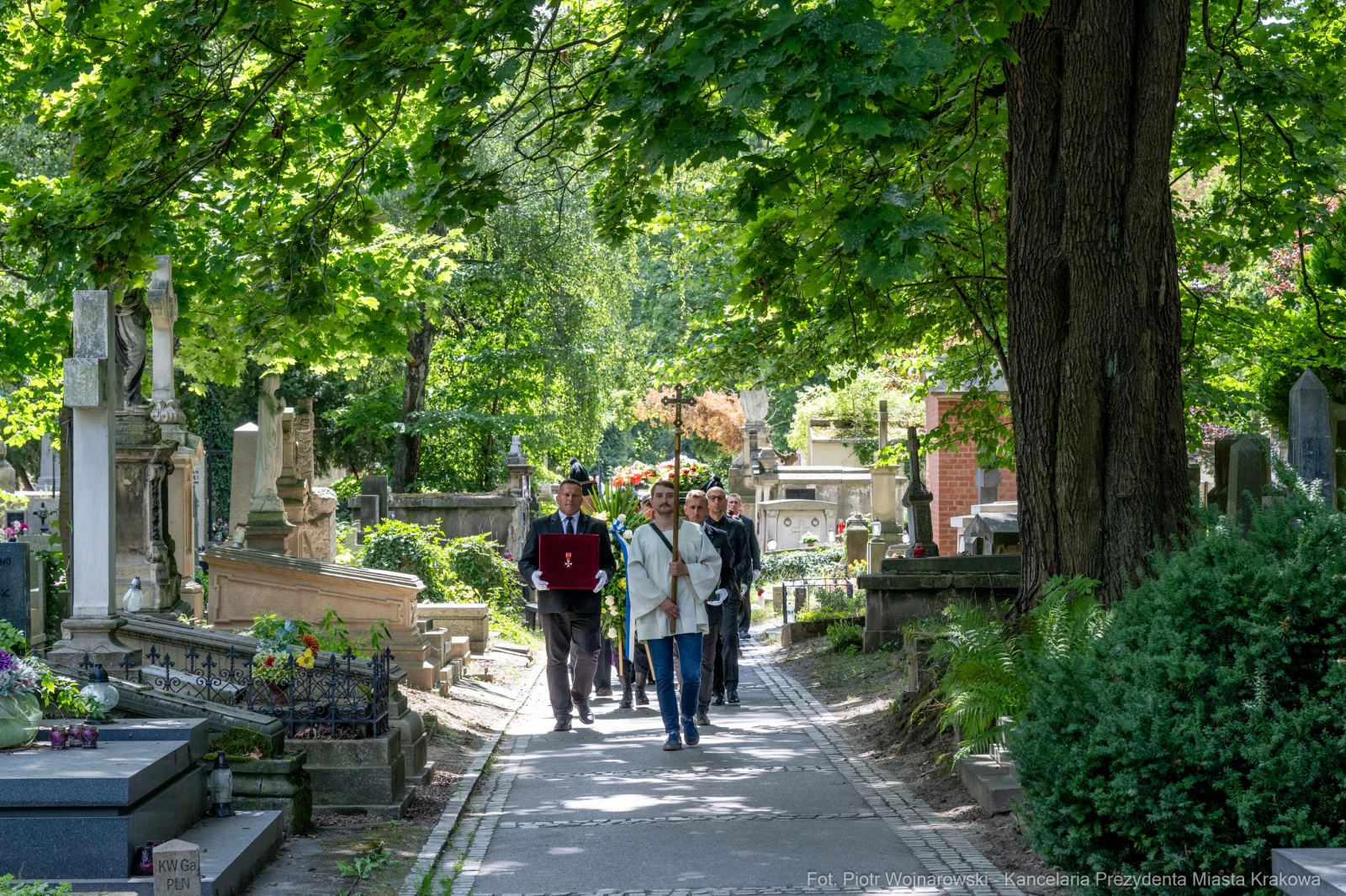Grupa ludzi spacerująca po cmentarzu w otoczeniu zieleni i nagrobków.  Autor: Piotr Wojnarowski