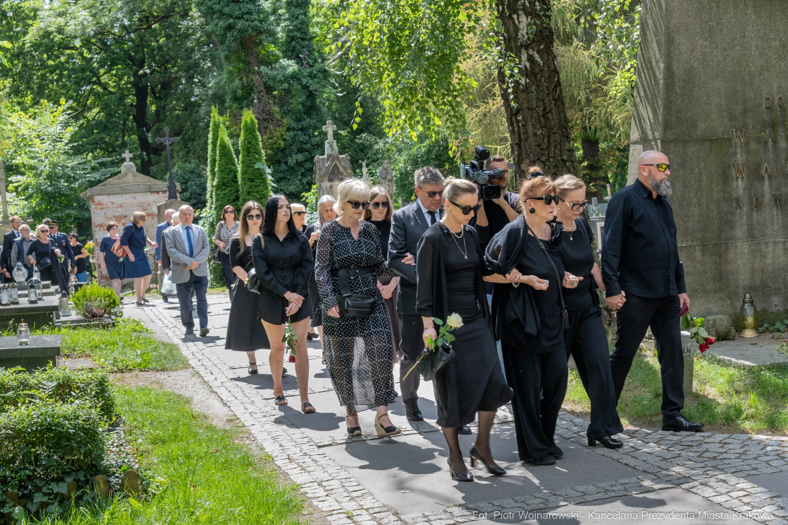 Grupa osób w czarnych strojach idzie alejką cmentarza, niosąc kwiaty.  Autor: Piotr Wojnarowski
