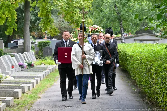Pochód żałobny z osobami niosącymi sztandar i zdjęcie zmarłego na cmentarzu.