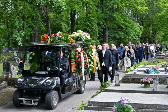 Pojazd z kwiatami prowadzi procesję pogrzebową na cmentarzu, uczestnicy w żałobie.
