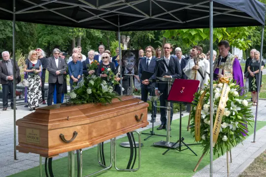 Pogrzeb z trumną, zebrani goście, kwiaty i przemawiający duchowny.