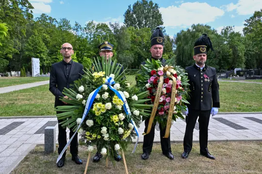 Czterech mężczyzn w mundurach stoi obok wieńców kwiatowych na cmentarzu.