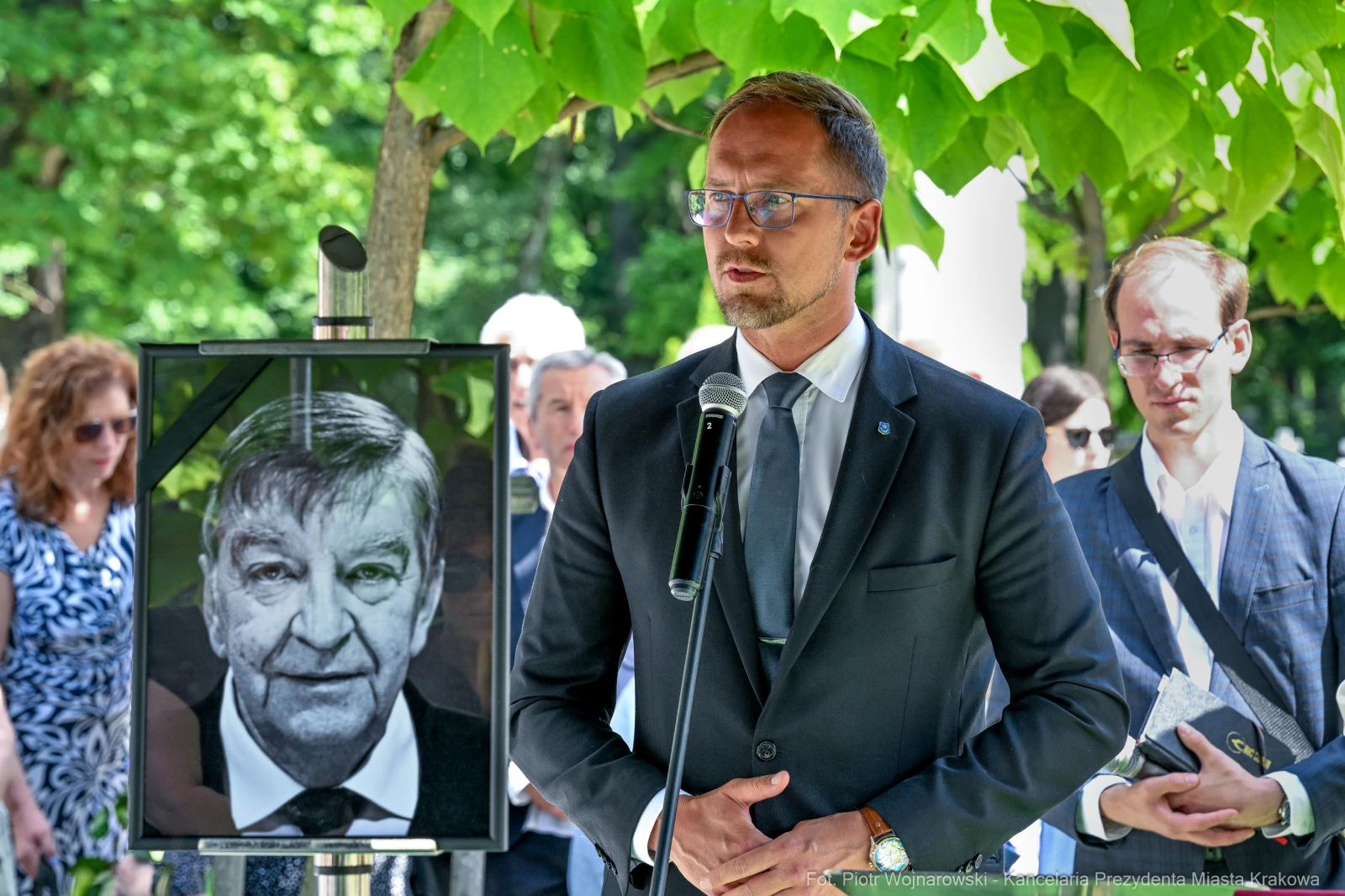Mężczyzna przemawia przy mikrofonie, obok portret w ramie. W tle zieleń i zgromadzeni ludzie.  Autor: Piotr Wojnarowski