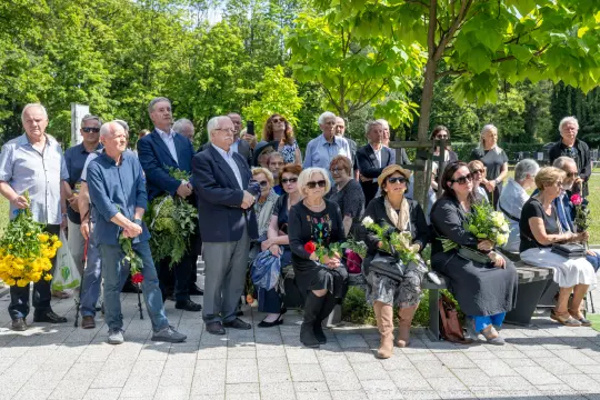 Grupa ludzi w różnym wieku, z kwiatami, uczestniczy w uroczystości na świeżym powietrzu.