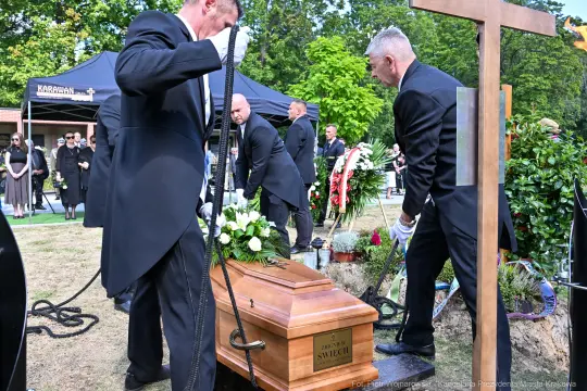Pogrzeb z trumną, mężczyźni w garniturach, kwiaty i krzyż w tle.