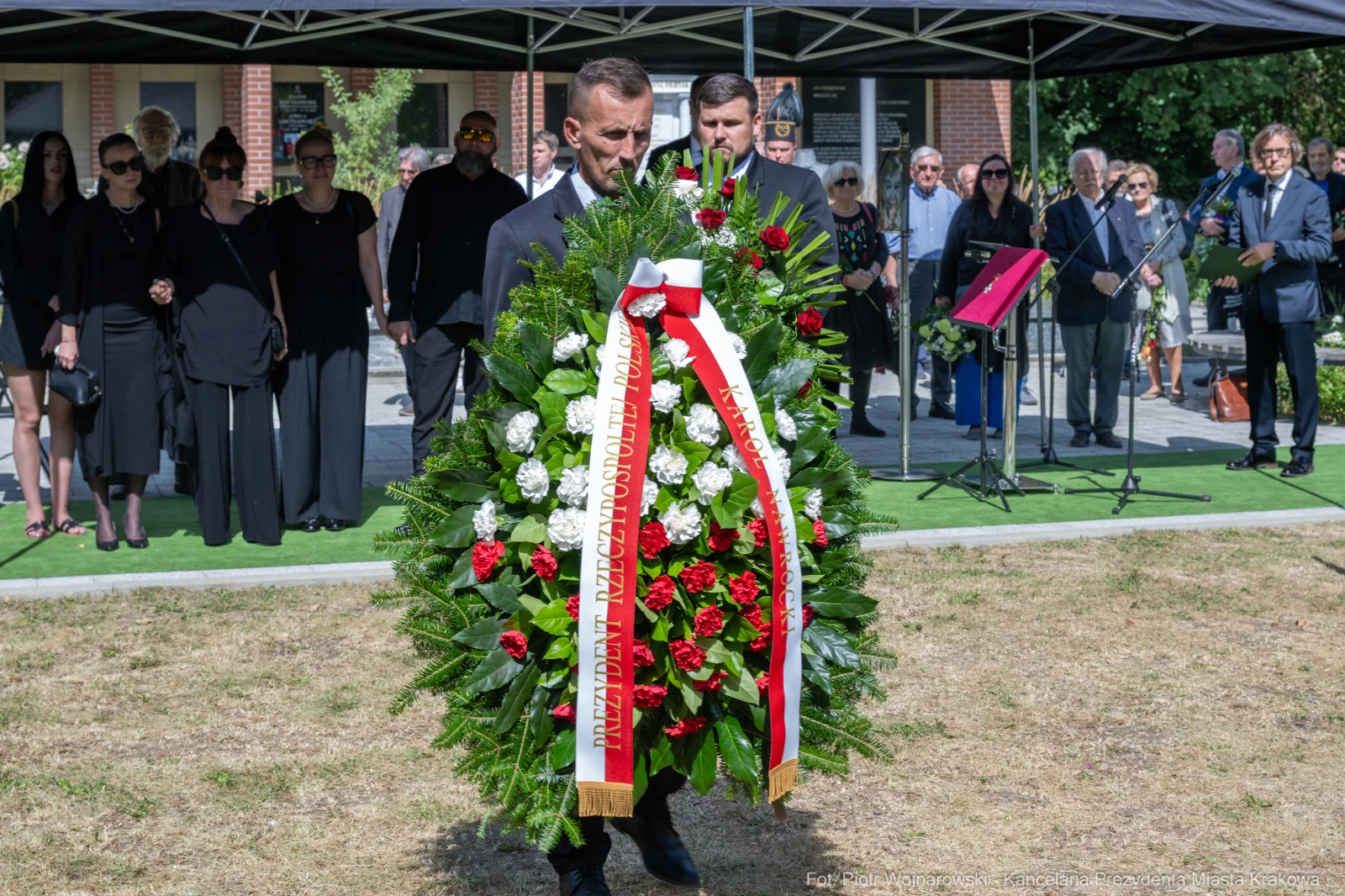 Mężczyzna niesie wieniec na ceremonii upamiętniającej, w tle zgromadzeni ludzie.  Autor: Piotr Wojnarowski