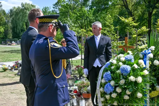 Ceremonia pogrzebowa z obecnością policjanta i mężczyzny w garniturze, kwiaty na grobie.