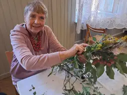 Starsza kobieta tworzy bukiet z kwiatów i liści, uśmiechając się.
