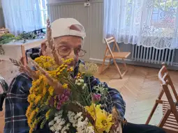 Starszy mężczyzna trzyma bukiet kolorowych kwiatów w pomieszczeniu z drewnianą podłogą.