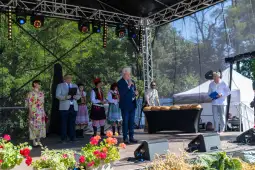 Uroczystość na scenie z mówcą i uczestnikami w tradycyjnych strojach. W tle stół z chlebem.