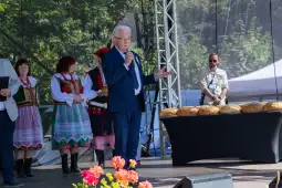 Mężczyzna w garniturze przemawia na scenie, obok niego osoby w tradycyjnych strojach ludowych.