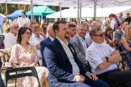 Grupa ludzi siedzących na wydarzeniu plenerowym, wśród nich mężczyzna w garniturze i kobieta w sukience.