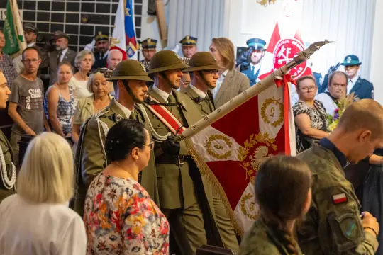 Ceremonia z udziałem żołnierzy niosących sztandar, tłum ludzi w tle.