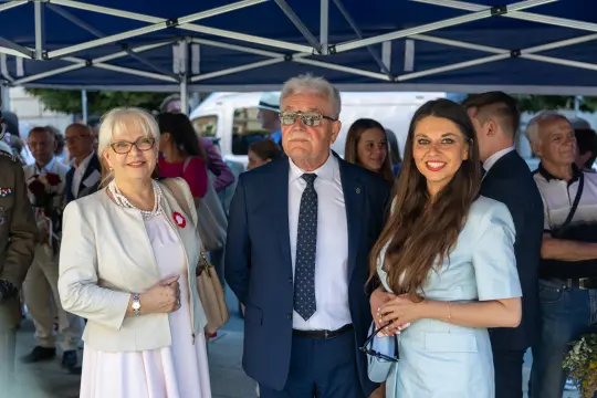 Trzy osoby w eleganckich strojach, uśmiechające się pod namiotem.