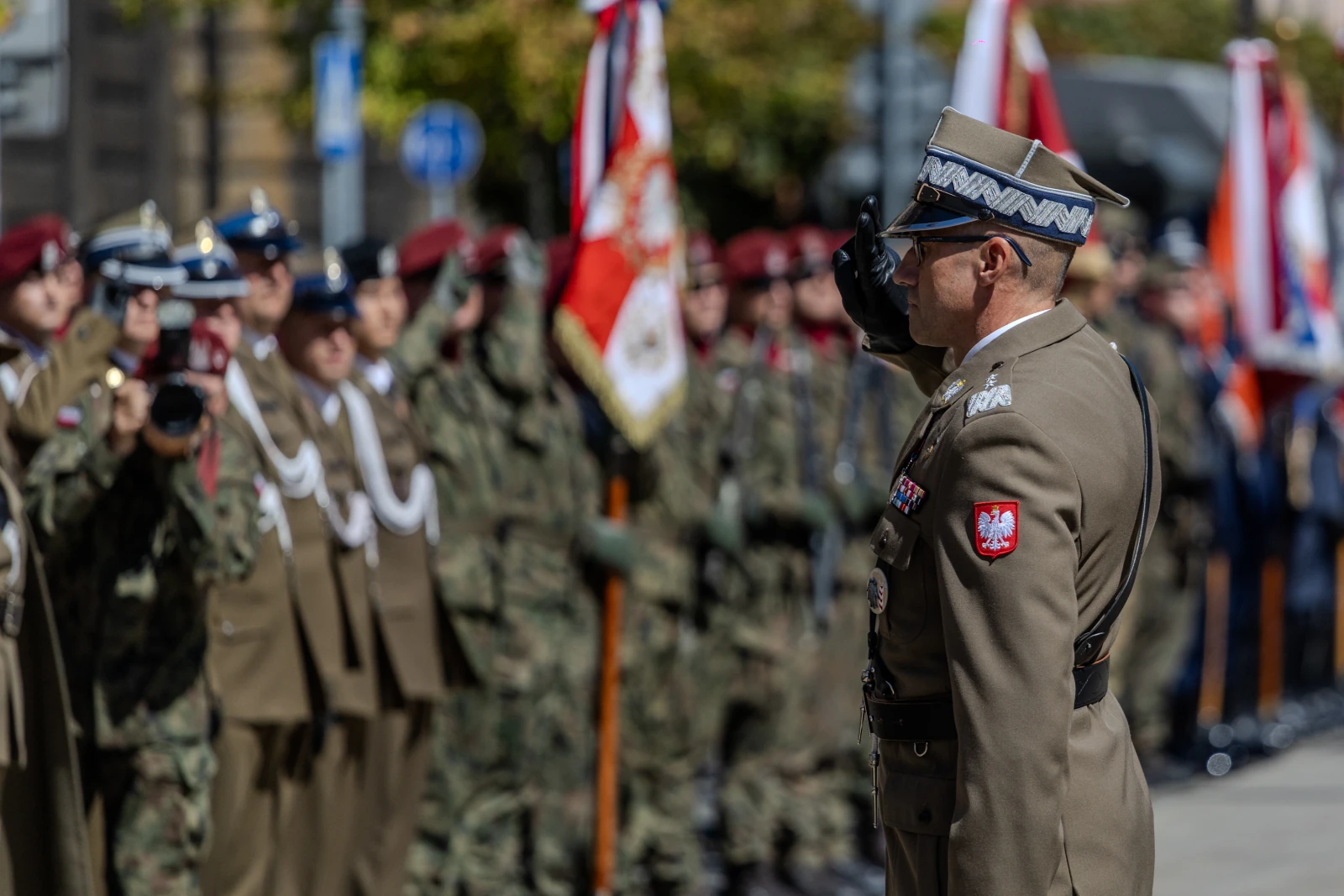 Żołnierz w mundurze wojskowym składa honory podczas uroczystości z flagami i żołnierzami w tle.  Autor: Bogusław Świerzowski