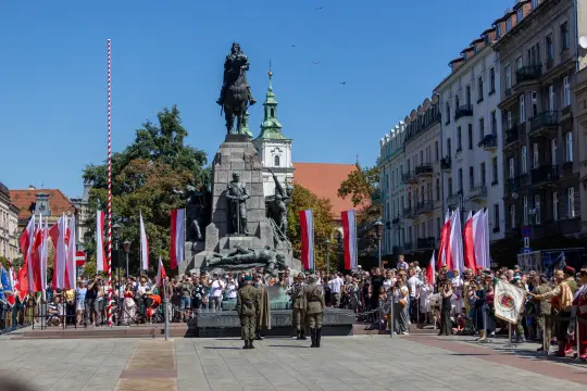 Uroczystość z udziałem żołnierzy i tłumu ludzi przy pomniku w centrum miasta. Flagi narodowe w tle.