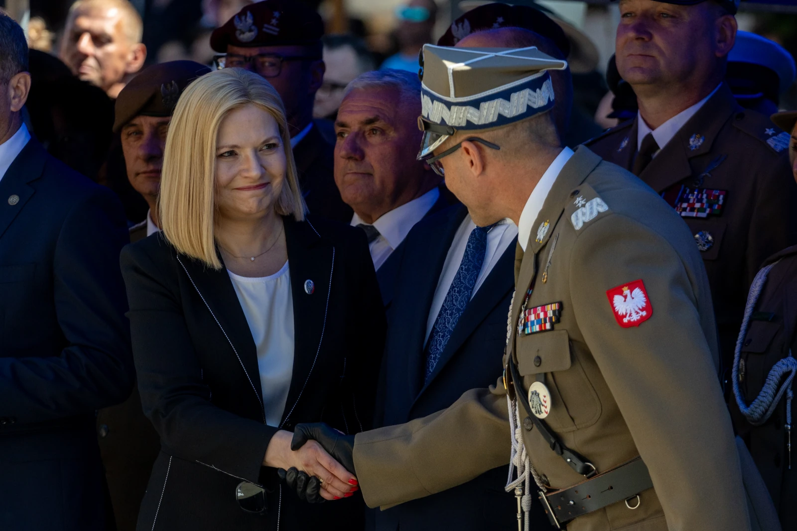 Uścisk dłoni między kobietą a mężczyzną w mundurze, w tle grupka mężczyzn w garniturach i mundurach.  Autor: Bogusław Świerzowski