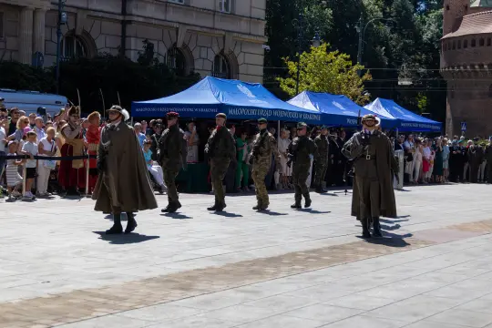 Żołnierze w mundurach maszerują przed tłumem ludzi, w tle namioty i budynki.