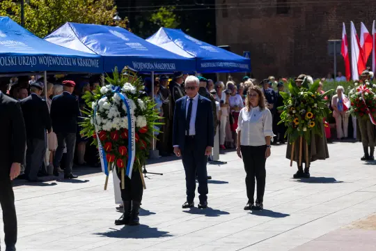 Ceremonia składania wieńców z udziałem oficjeli i uczestników, flagi w tle.