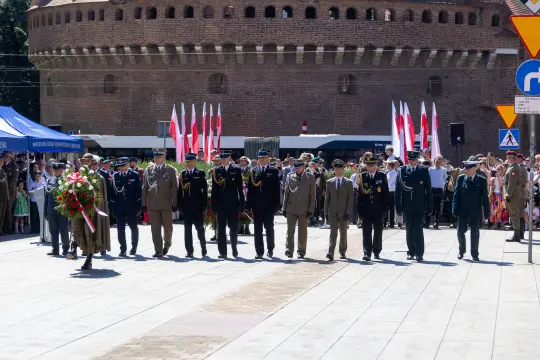 Uroczystość z udziałem wojskowych i flag w tle, z historycznym budynkiem w tle.