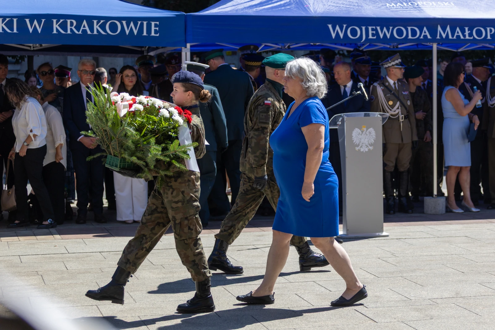 Kobieta w niebieskiej sukience idzie obok żołnierza niosącego kwiaty podczas uroczystości.  Autor: Bogusław Świerzowski