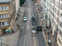 Tramwaj na ulicy, obok samochody i piesi, budynki w tle.