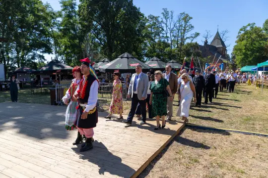 Grupa ludzi w tradycyjnych strojach idzie w kierunku sceny podczas wydarzenia plenerowego.