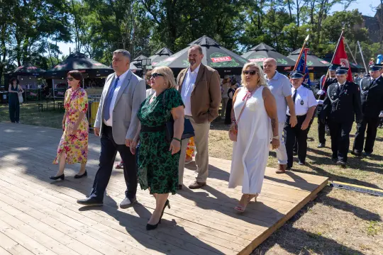 Grupa osób w eleganckich strojach idzie po drewnianym podłożu podczas wydarzenia plenerowego.