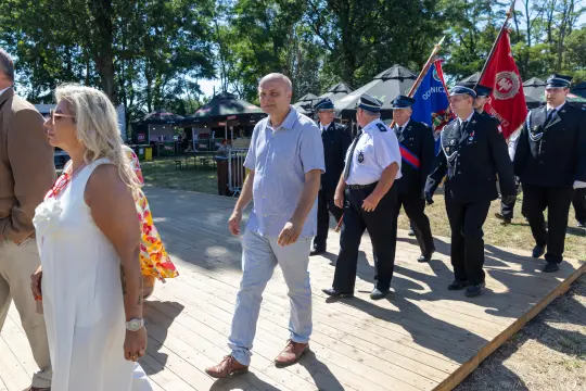 Grupa ludzi w mundurach i cywilnych ubraniach idzie po drewnianym podeście w otoczeniu drzew.