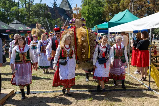 Grupa osób w tradycyjnych strojach ludowych podczas festiwalu, obok stoiska i namiotów.